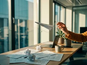 Papierflieger wird von Hand sanft umgeleitet über modernem Bürotisch mit zerknüllten Skizzen bei warmem Tageslicht